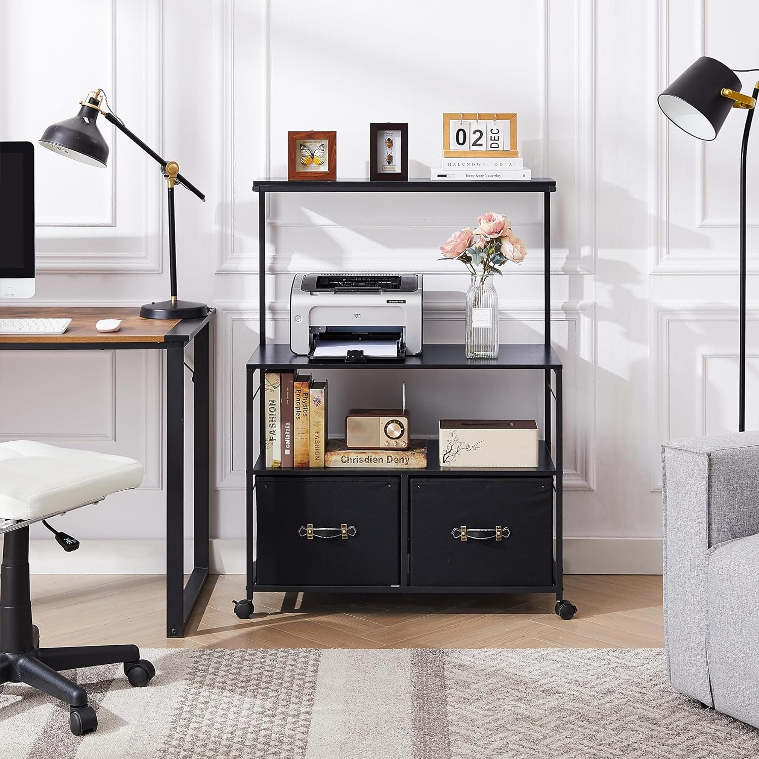 Modern black printer stand with shelves and storage drawers, placed beside a computer desk in a bright home office, holding a printer, books, and decorative items.