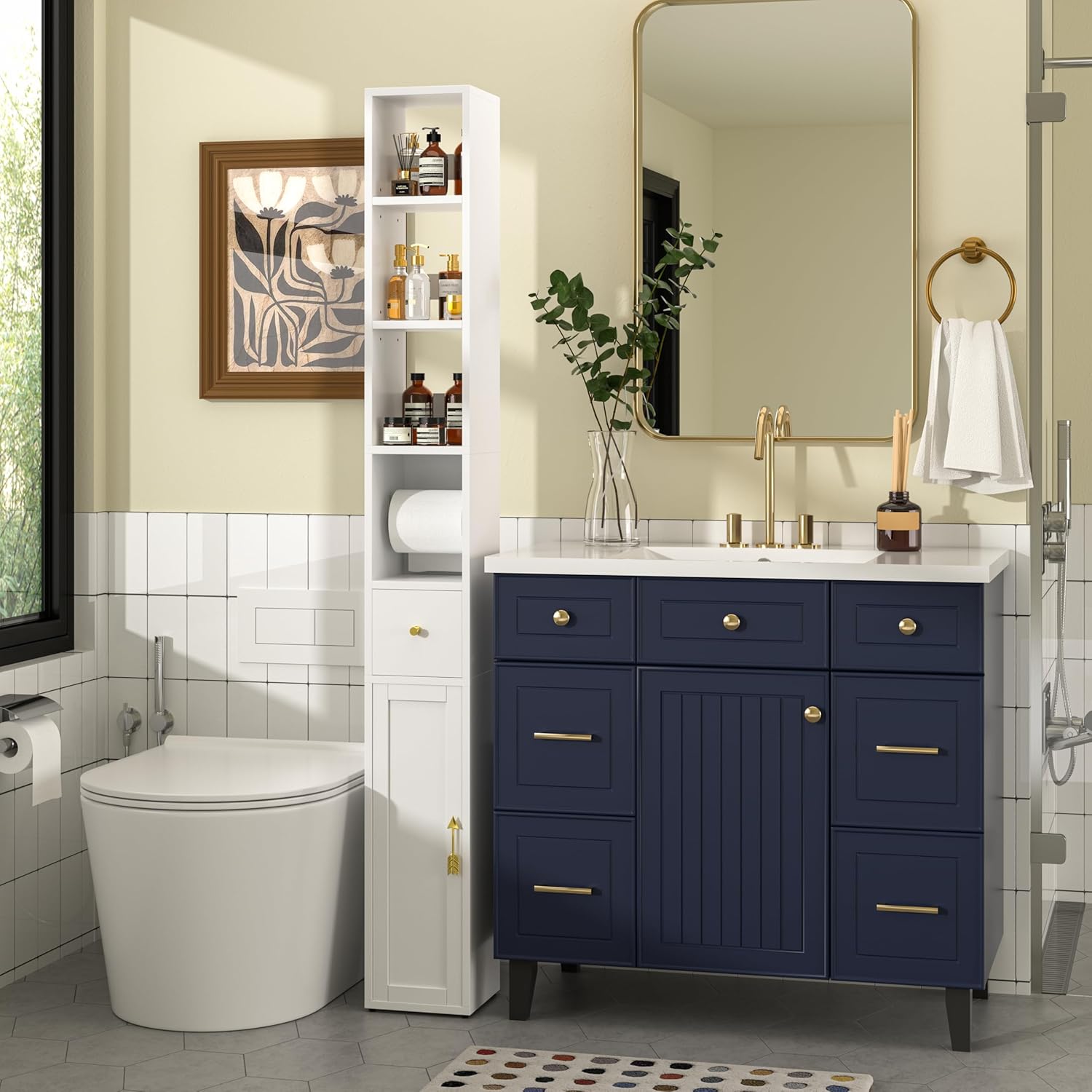 Tall narrow white bathroom cabinet beside navy blue vanity with gold hardware, used for organized storage of toiletries and toilet paper.